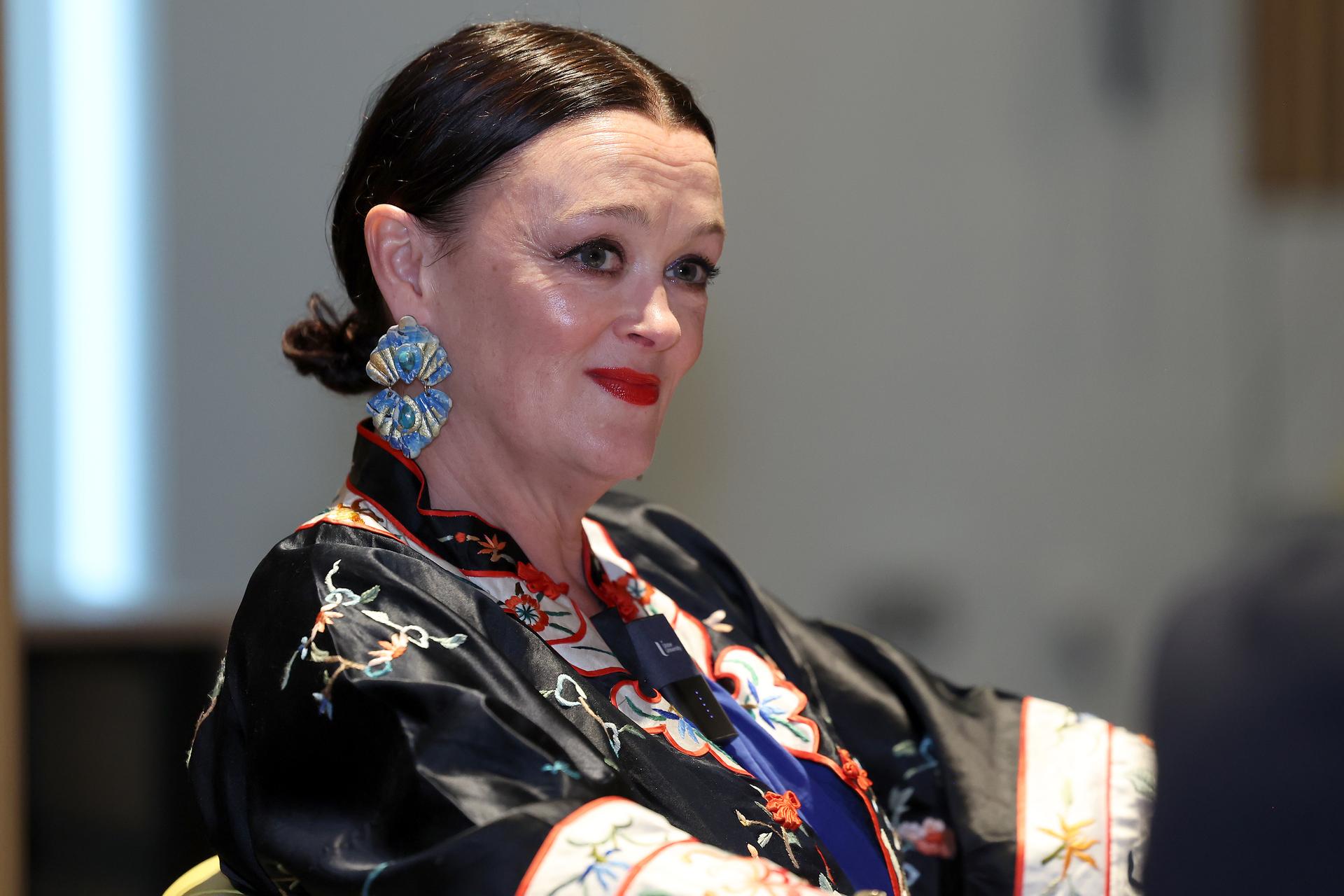 Close-up of a speaker seated during a discussion event, wearing a black embroidered jacket with colourful floral detailing and large blue statement earrings. A small microphone is clipped to the jacket. The background is softly blurred, keeping the focus on the speaker.